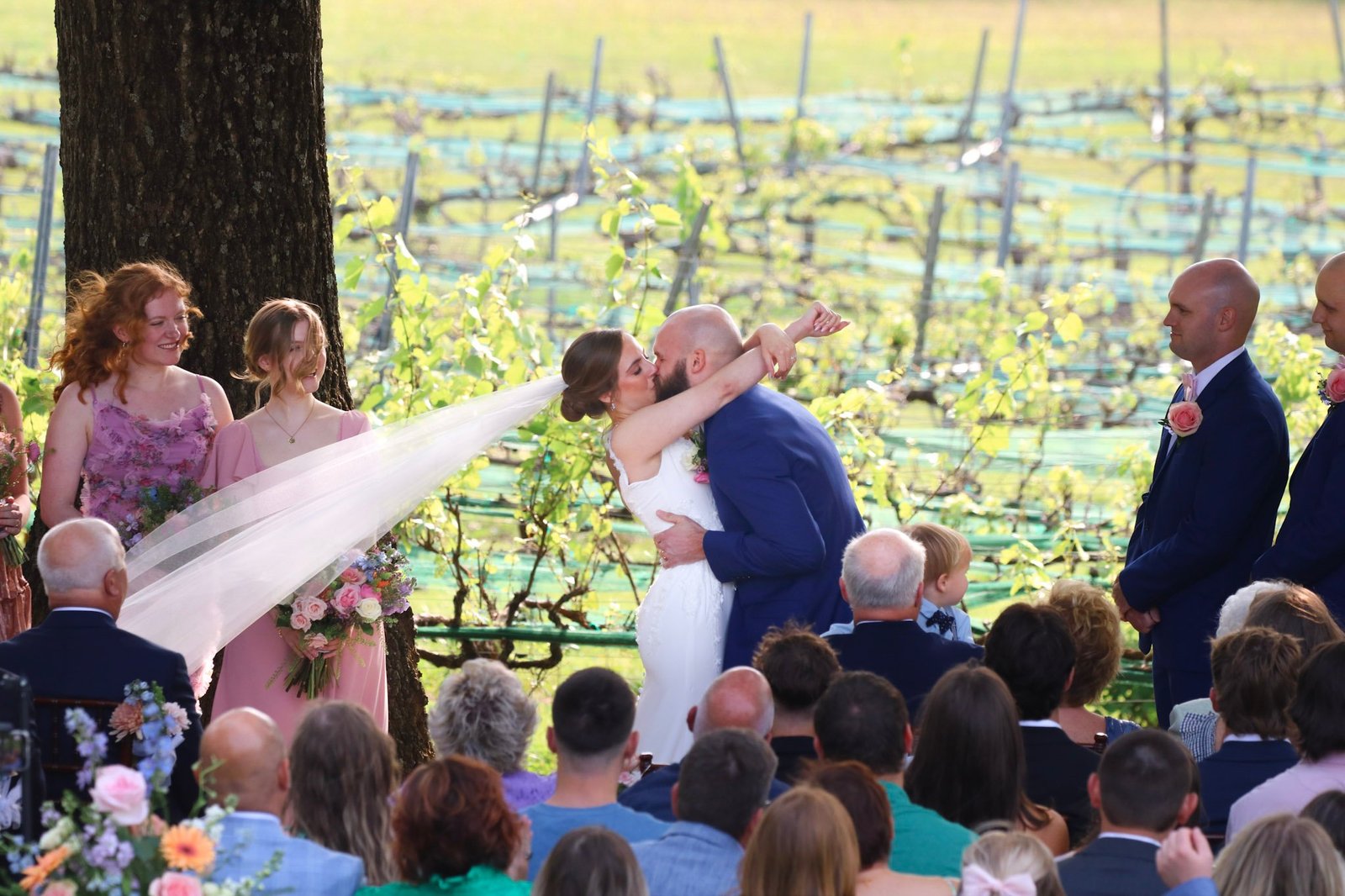 Bride and groom share a first kiss during vineyard ceremony with bridesmaids and groomsmen looking on at Pilot Mountain Vineyards