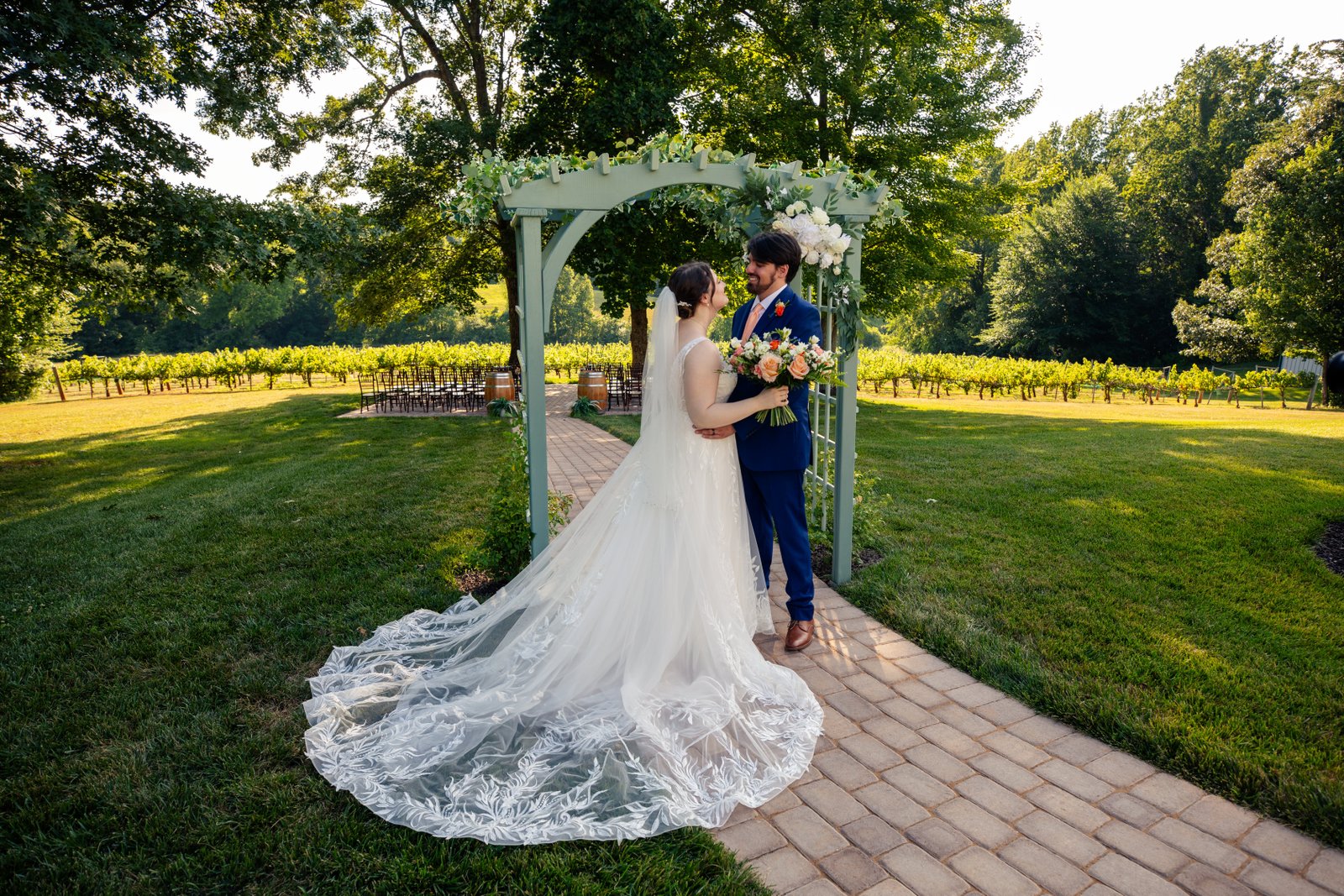 Bride and groom exchange vows under a floral archway on brick pathway with long train flowing and vineyard behind at Pilot Mountain Vineyards