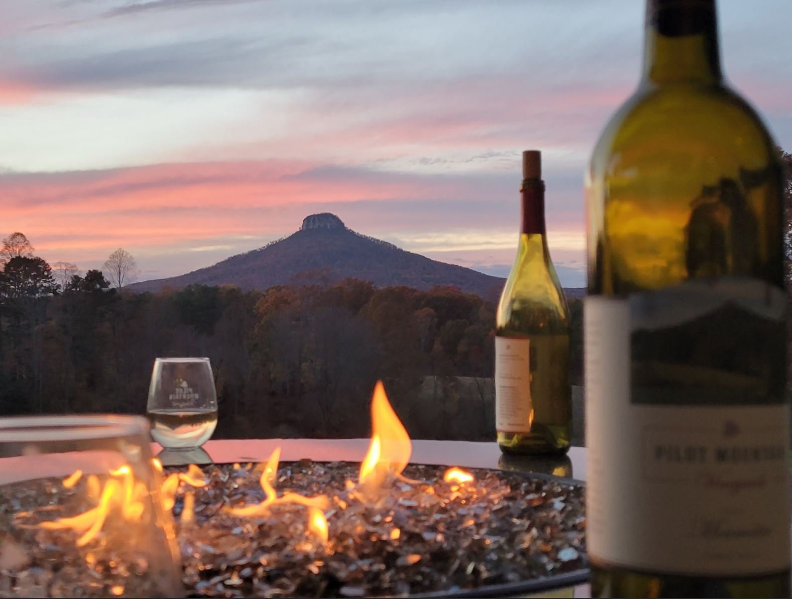 Wine bottle and glass near fire pit with flames reflecting, overlooking Pilot Mountain at sunset