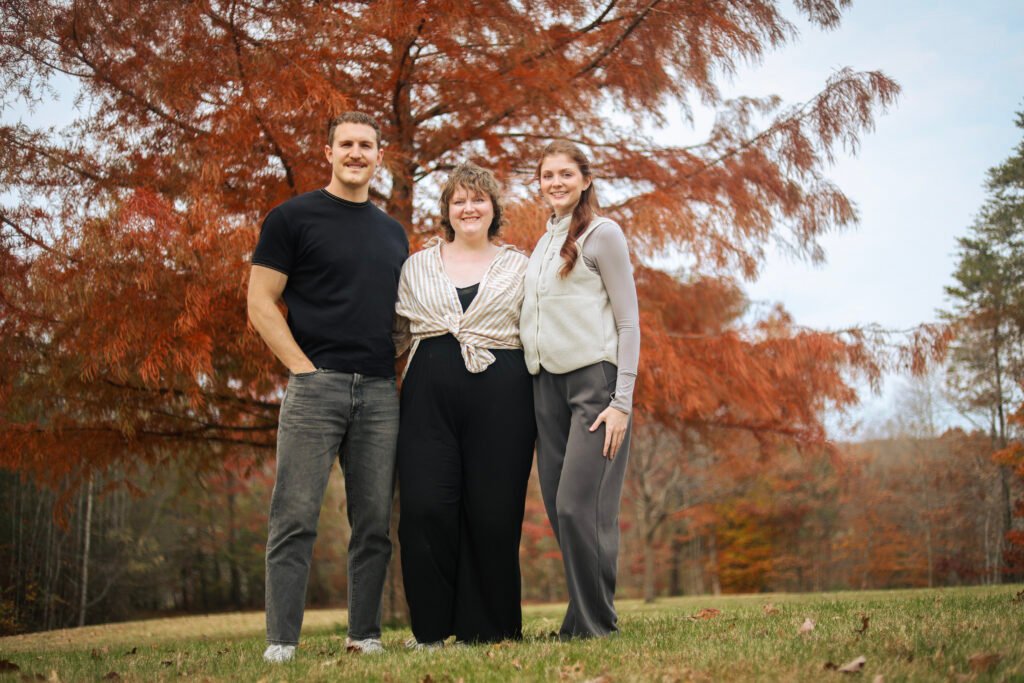One man and two women posing and smiling in front of a big red tree